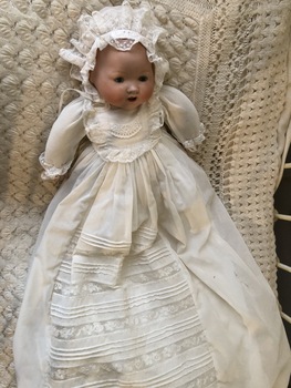 A porcelain or ceramic baby doll dressed in a long white cotton and lace dress laying on a white crocheted rug.