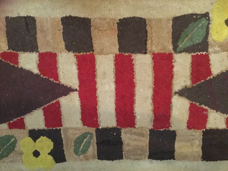 A small handmade highly patterned wool floor rug with red and brown stripes as well as green leaves and yellow flowers.