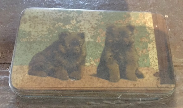 A green and brown painted chocolate tin with two dogs pictured on the lid.