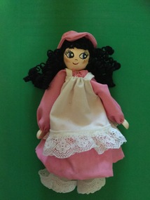 A peg doll dressed in a pink dress and hat with a white apron and pantaloons.