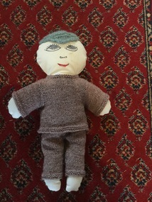 A white fabric handmade doll with a painted face, a blue knitted hat and brown clothes.
