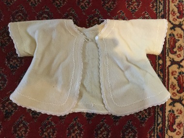 A cream woollen baby matinee jacket with herringbone stitching and a button and loop to close it at the neck.