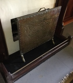 A brass firescreen with a raised design on the front, a handle on the top and is footed.