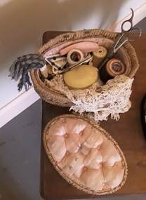 A cane and fabric covered sewing basket with assorted items for domestic sewing in the home.