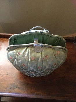 A large pale green satin padded tea cosy with flowers and triangles on the sides. It has a clip at the top to close it.