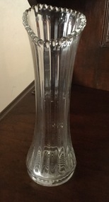 A tall clear glass vase with a vertical linear pattern and fluted edging.