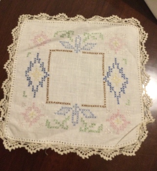 A small square cotton embroidered doily with brown, green, blue, pink, white and navy cross stitch pattern. 