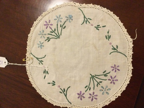 A small round floral embroidered doily in blue, purple and green thread with a scalloped edge. 