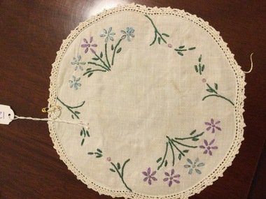 A small round floral embroidered doily in blue, purple and green thread with a scalloped edge. 