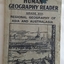 Grey paperback Whitcomb'e Human Geography Reader for Grade VIII with a picture of a city next to a river with wharf activity on the opposite side of the river. The cover is worn and ink stained with Lance Sebire written at the top.