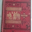 A red covered book with gold imprint of a picture of schoolboys at a desk in a classroom. The title Arthur's Victory and author A. E. Ward are printed in red surrounded by gold boxes with black fleur de lis patterns over the whole front cover. The book was published in 1873 so shows signs of wear but is in reasonable condition for its age.