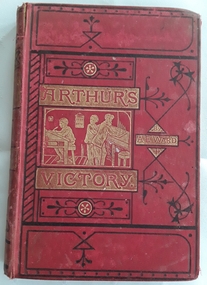 A red covered book with gold imprint of a picture of schoolboys at a desk in a classroom. The title Arthur's Victory and author A. E. Ward are printed in red surrounded by gold boxes with black fleur de lis patterns over the whole front cover. The book was published in 1873 so shows signs of wear but is in reasonable condition for its age.