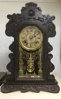 A very ornately carved dark wooden mantel pendulum clock with a white face and a highly decorated glass front.