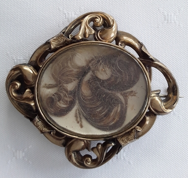 A swirled patterned highly decorated brass brooch with human hair under a glass cover.