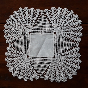 A square cotton handmade scallop edged doiley for use on furniture to protect the surface.