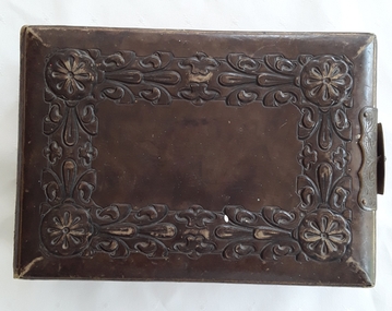 An antique thick rectangular photograph album of dark tan leather with an elaborate embossed floral and leaf design on the covers.