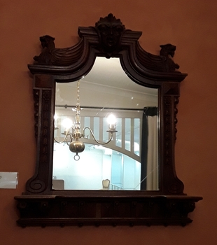 An ornately hand carved rectangular wooden framed mirror with a shelf at the bottom and a carved head at the top.