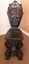 An ornately hand carved dark wooden heavy nursing chair with a carved figure of a child and gargoyles on the front of the back rest.