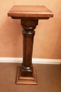 A tall wooden pedestal with a square top, a column with a vertical lined pattern, mounted on a two tiered base.