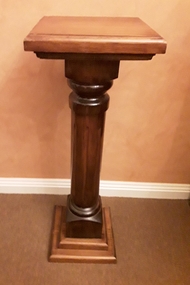A tall wooden pedestal with a square top, a column with a vertical lined pattern, mounted on a two tiered base.