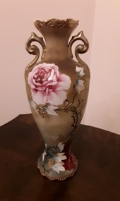 A tall slim gold vase with two handles decorated with pink roses and gold embossed leaves.