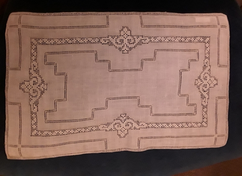A rectangular ornamental machine made doiley made of white cotton with a cut out pattern.