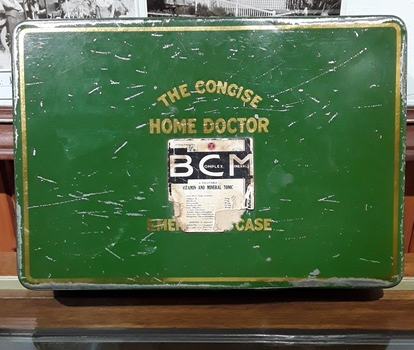 A dark green lidded tin with gold trim around the edges and gold lettering stating 'The Concise Home Doctor Emergency Care'. 