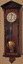 A tall narrow pendulum wooden wall clock with a white round clock face, with Roman numerals, elaborate fine hands and a brass pendulum.