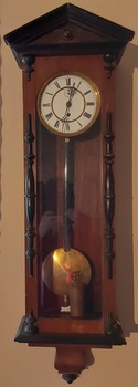 A tall narrow pendulum wooden wall clock with a white round clock face, with Roman numerals, elaborate fine hands and a brass pendulum.