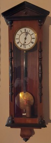 A tall narrow pendulum wooden wall clock with a white round clock face, with Roman numerals, elaborate fine hands and a brass pendulum.