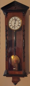 A tall narrow pendulum wooden wall clock with a white round clock face, with Roman numerals, elaborate fine hands and a brass pendulum.