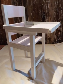 A white and lilac painted wooden homemade dolls high chair with a lift up tray, four legs, two arms and a back support.