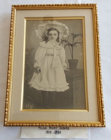 A framed photograph of Hilda Rihoy Sebire 1899 - 1906, taken standing next to a decorative stand with a pot plant fern.