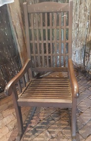 A dark brown wooden rocking chair with two shaped arms which have three wooden supports  on each side.