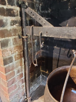 A black cast iron fireplace crane with a supporting pole bolted to the wall in the side of the brick open fireplace. 