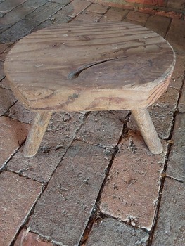 A replica of a hand hewn wooden milking stool which is round with two flat sides with three round legs glued underneath. 