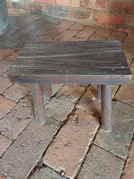 A replica of a rough hand hewn rectangular hardwood milking stool with four round wooden legs glued into holes underneath the seat.