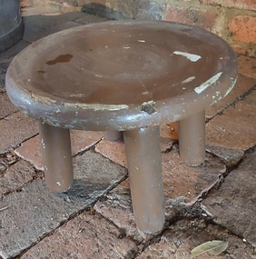 A round brown painted wooden milking stool with four round wooden legs glued into holes underneath the seat.