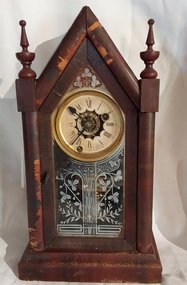 A small vintage decoratively carved dark timber steeple mantel pendulum clock with a white cardboard face, black roman numerals and black metal hands.