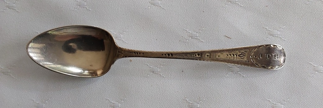 A single silver teaspoon with engraving on the top of the handle and a maker's mark underneath.