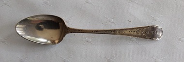 A single silver teaspoon with engraving on the top of the handle and a maker's mark underneath.