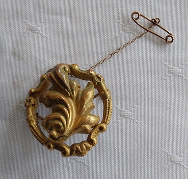 A round gold decorative filigree brooch with a central large leaf in the middle of a circular shape with a tiny safety chain. 