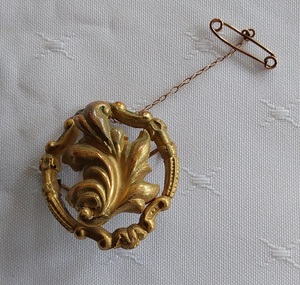 A round gold decorative filigree brooch with a central large leaf in the middle of a circular shape with a tiny safety chain. 