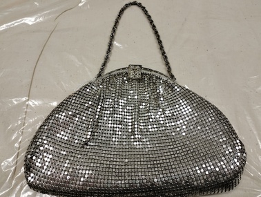 Front of small silver purse, half moon shaped with small chain handle. Ruched at top to a small frame opening closed by a square metal clasp. Made of silver mesh chain mail.