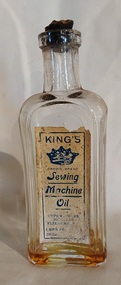 A clear glass narrow rectangular sewing machine oil bottle with a short neck and a broken cork stopper. 