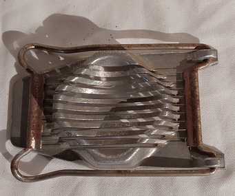 A stainless steel egg slicer with a base which has a raised stand or support with slats to hold the cooked, shelled egg.
