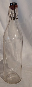 A large clear glass Fowlers Fruit Juice bottle with a wire clip and black stopper with a red rubber seal. 