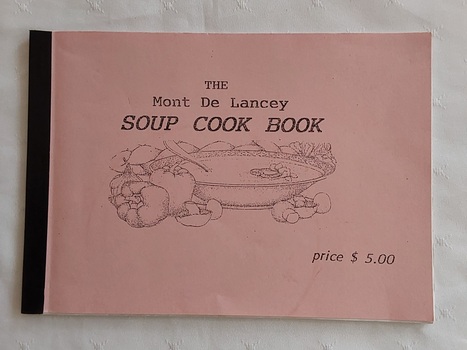 A small rectangular pink covered home made Soup Cook Book with the title and an illustration of a bowl of soup with vegetables around it.