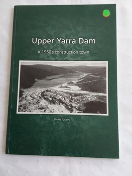 A green covered paperback book with the title printed in white, a black and white aerial photograph of the town and dam, and the author below. 