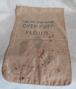 A vintage small cream coloured draw string cotton flour bag with the brand Oven-Puff Flour printed in faded pale blue lettering on both sides.